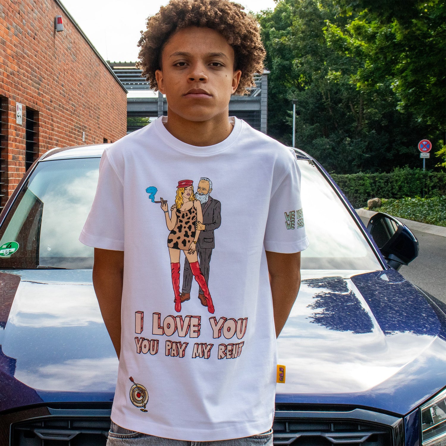 Model wearing Allmosst white t-shirt with graphic design and text, standing in front of a car.