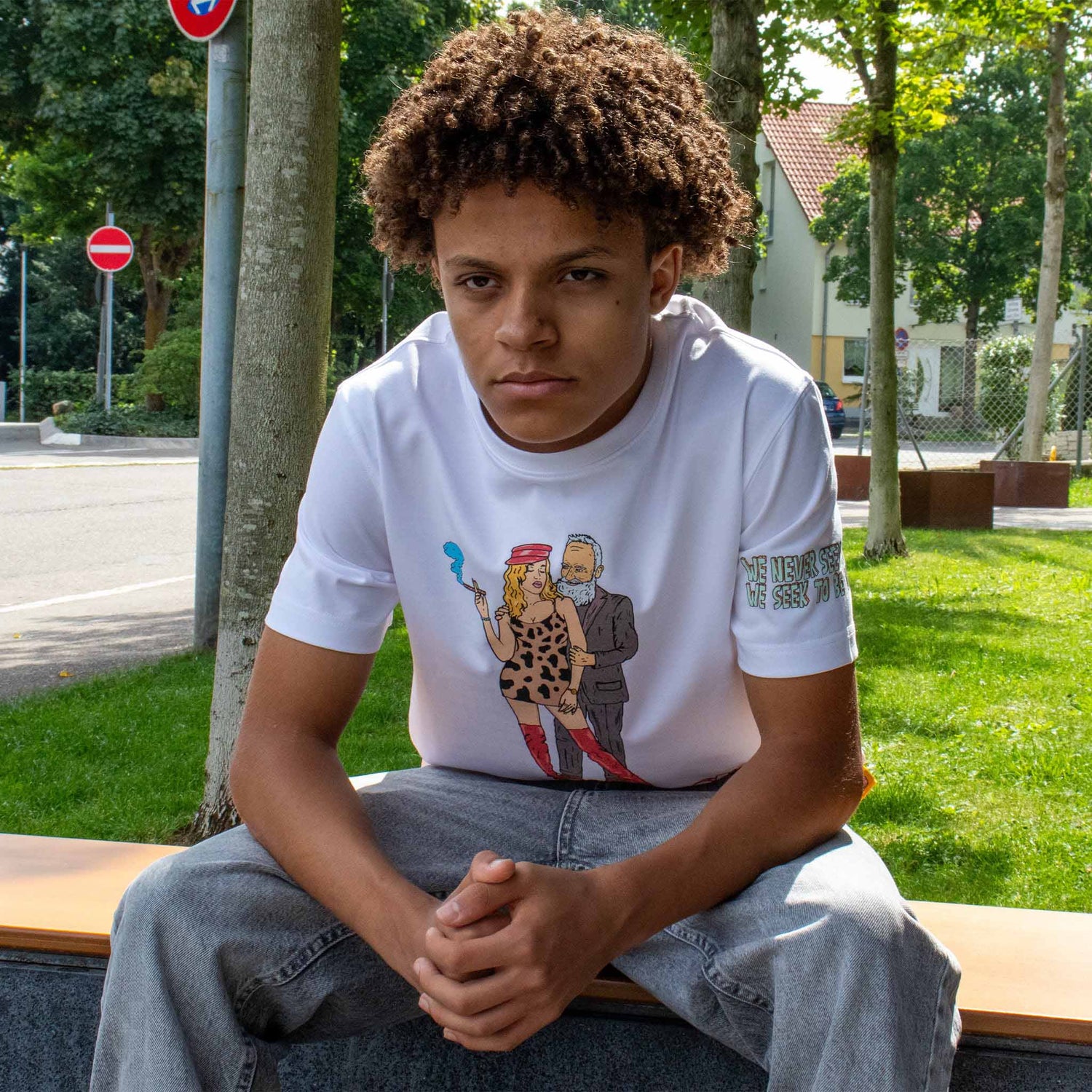Model wearing Allmosst white t-shirt with graphic design sitting on a bench outdoors.